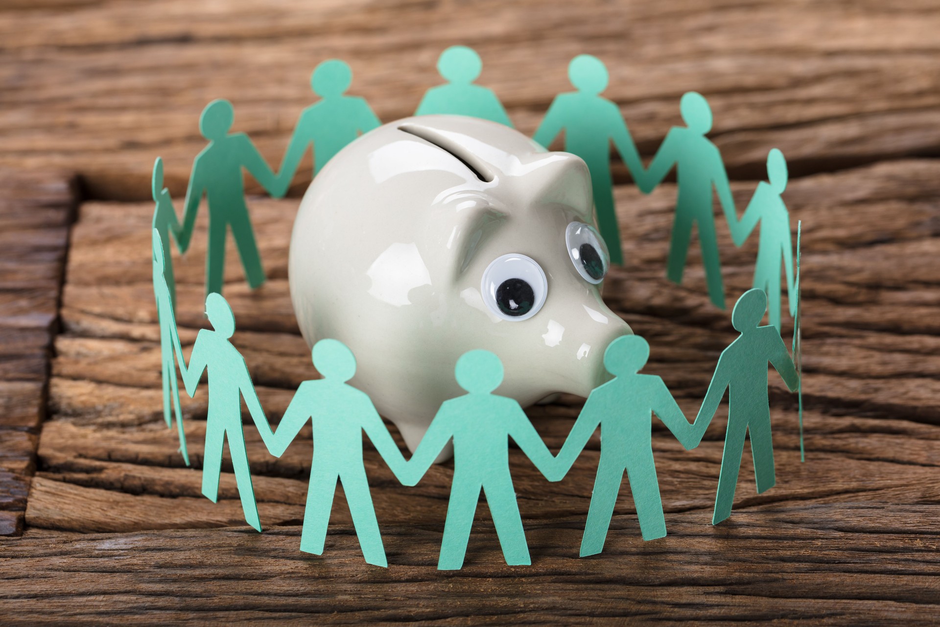 Piggybank Surrounded By Paper People Holding Hands On Wood